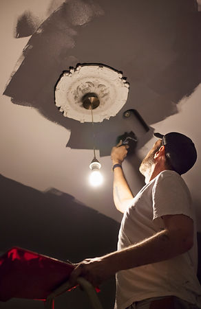 Man wearing a cap paints a ceiling near an ornate light fixture with a roller
