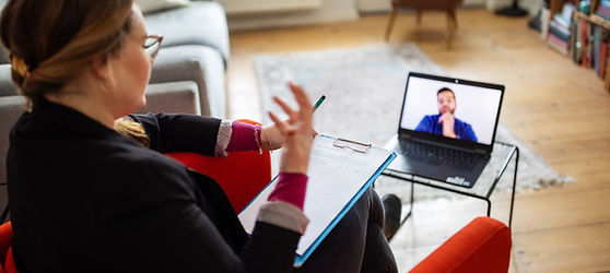 Black woman wearing glasses sitting in a red chair with clipboard on her lap speaking with a man on a laptop