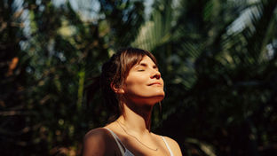 Woman in white top stands in sunlight with eyes closed, smiling contentedly. Lush green foliage in background creates a peaceful ambiance.
