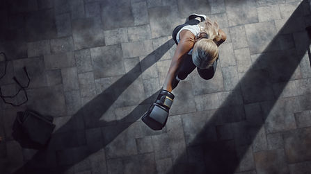 It's an aerial overhead picture of a woman with long blonde hair. She is located on a gray tile floor. She is wearing a white tank top, black-and-white shoes, and black joggers. She is throwing a punch, with boxing gloves on.