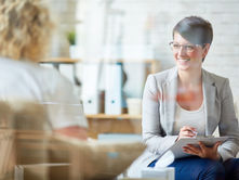 Smiling woman therapist in glasses with clipboard converses with another person in a bright office. Background includes blurred bookshelves.