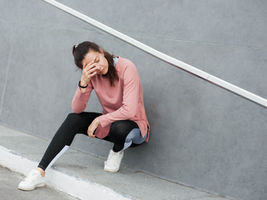 Woman in pink top and black leggings squats against gray wall, hand on forehead, appearing exhausted. White sneakers, concrete background.
