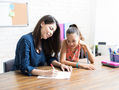 A female teacher helps a girl write at a wooden table in a bright classroom, with a whiteboard and school supplies in the background.