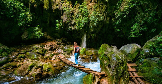 Woman walks across a wooden bridge over a river in lush forest.