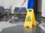 Person mopping a shiny office floor with blue chairs. Yellow "wet floor" sign in foreground. Calm setting with a potted plant.