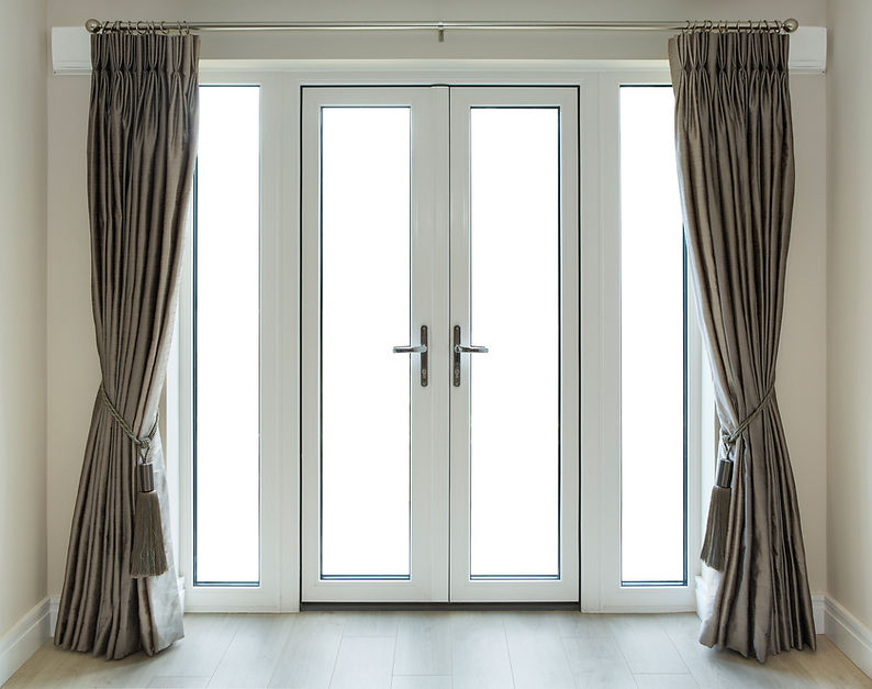 A set of closed white French doors flanked by tall, thin windows, adorned with elegant, tied-back taupe curtains
