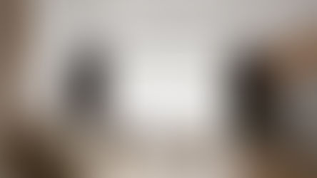 Modern living room with beige sofa, cushions, and pouf on wooden floor. Large window with sheer curtains, TV, and indoor plants. Calm ambiance.