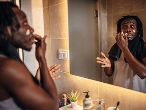 Man applying face cream in bathroom, looking in mirror. Towel draped over shoulder, tan wall background, small plant by sink. Reflective mood.