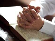 Clasped hands in prayer over an open Bible on a wooden table, warm sunlight creating a quiet, reverent mood.