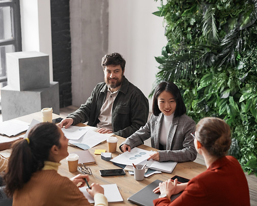 Five people in a meeting at a wooden table with papers and laptops, a leafy green wall backdrop, and large windows. They appe