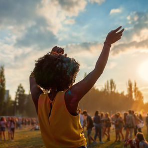 Person in a yellow tank top with arms raised enjoys a sunny outdoor festival. People gather in the background, creating a joyful atmosphere.