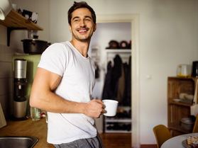A smiling man in a white T-shirt holds a coffee mug in a cozy kitchen. Shelves, kitchenware, and a small table are visible in the background.