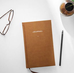 A brown journal, glasses, a pen and coffee sit on a desk
