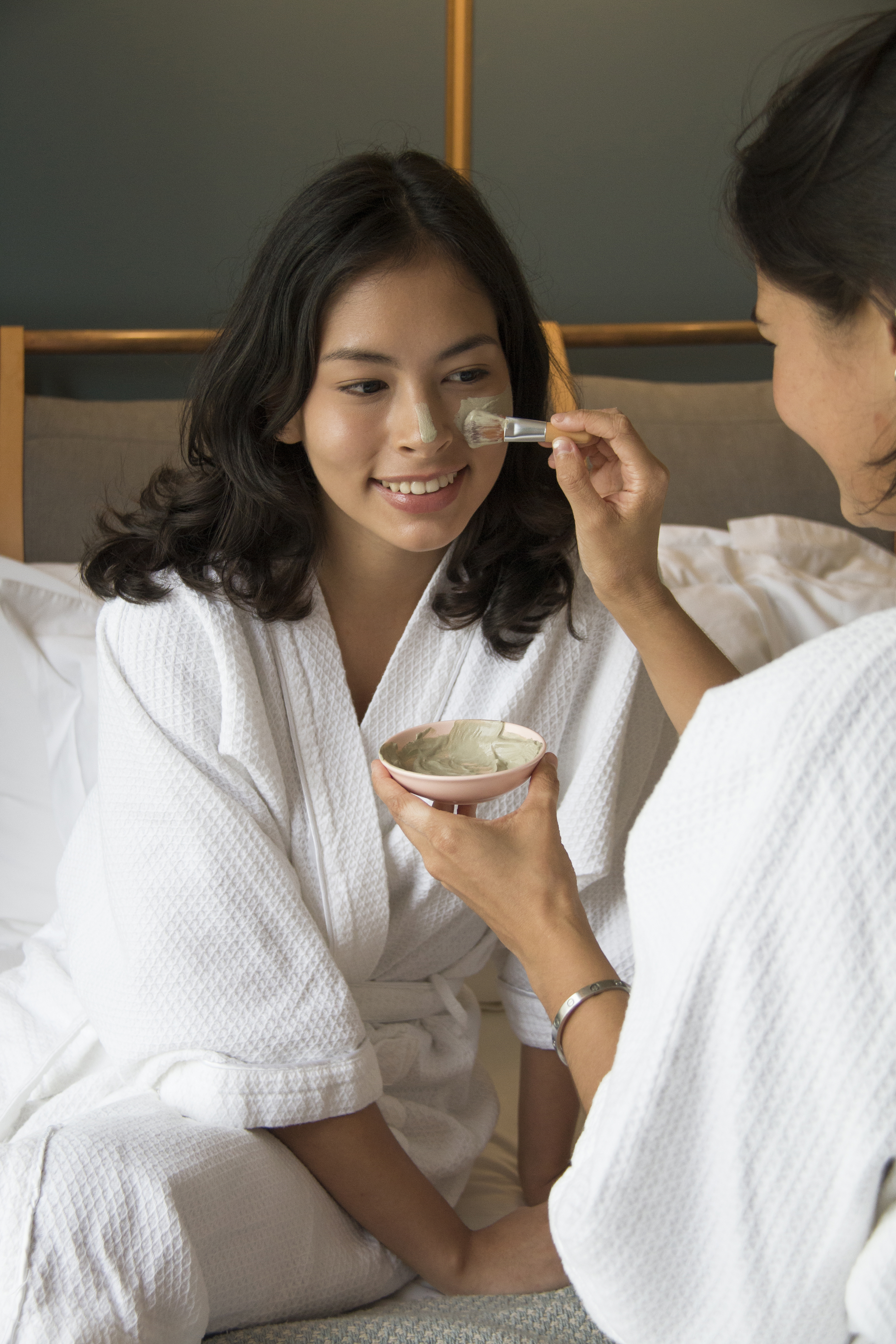 Two women in white robes, one applying a facial mask to the other in a cozy indoor setting.
