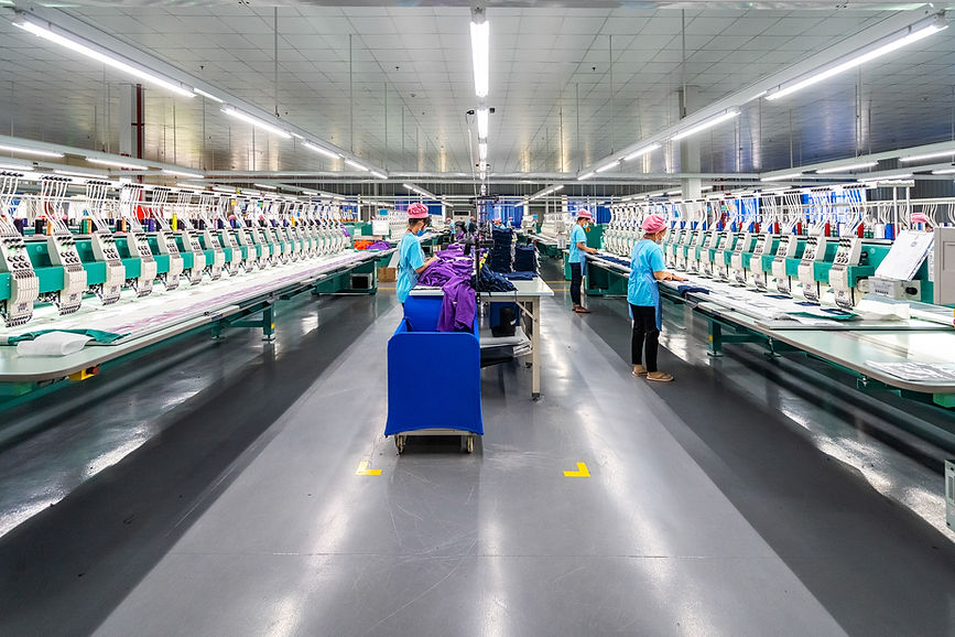 A brightly lit factory floor features rows of embroidery machines with workers in blue uniforms