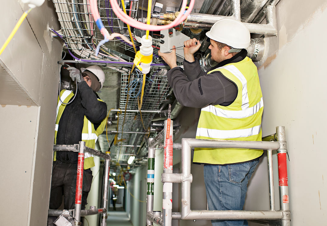 Two construction workers in safety vests and helmets work on ceiling wiring in a narrow corridor