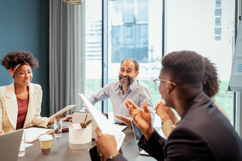 Colleagues in a meeting room discuss documents, smiling. Modern office with large windows, bright and collaborative atmosphere.