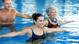 Three people smiling while doing water exercises in a pool. Clear blue water, bright swimsuits, and cheerful mood.