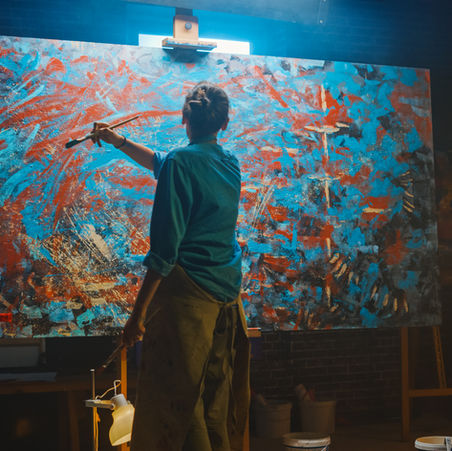 Artista em pé diante de uma grande tela abstrata, pintando com pincel gestos largos em tons de azul, vermelho e laranja. A cena acontece em um ateliê com luz direcionada sobre a obra, revelando textura espessa de tinta, movimento do corpo e concentração no processo de criação.