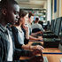 Students work at desktop computers in a library lab, with a focused young man in plaid in the foreground.