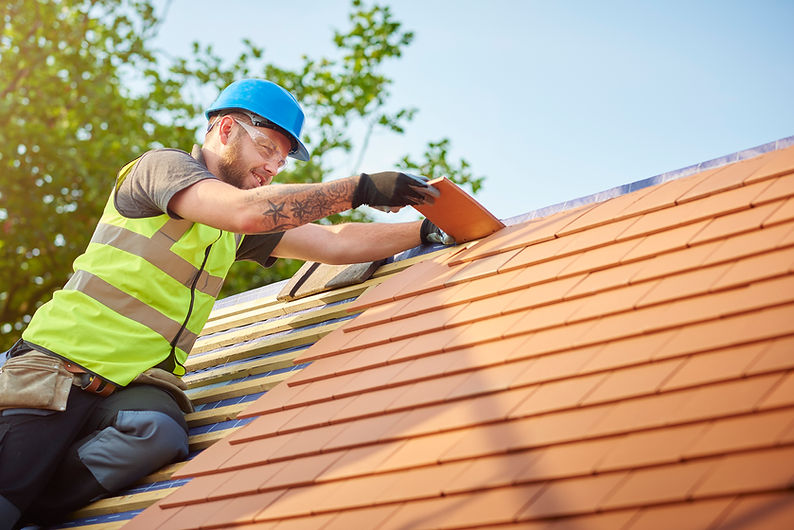 A construction worker in a bright vest and blue helmet installs orange tiles on a roof