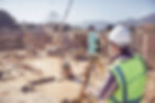 A construction worker in a helmet and vest uses surveying equipment on a busy construction site