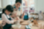 A woman and child in aprons mix batter in a bowl, smiling in a cozy kitchen. Sunlight streams through a window onto a wooden counter.