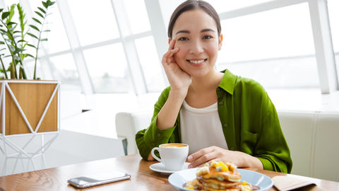 Femme souriante dans un café lumineux, veste verte, devant un café, des pancakes et un smartphone sur la table.
