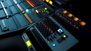 Close-up of a dimly lit audio mixing console with colorful LED lights and glowing meters, indicating an active recording studio setting.