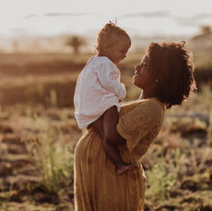 a pregnant mother standing outside holding her baby sitting on top of her baby bump. Mom and baby are looking at each other and smiling.