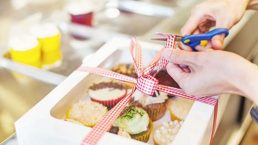 Hands cut a red checkered ribbon on a white bakery box filled with cupcakes. Background includes blurred yellow and red cupcake liners.