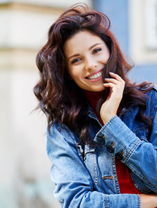 Smiling woman in a denim jacket poses outdoors against a blue building. She gently touches her cheek, creating a joyful, relaxed mood.