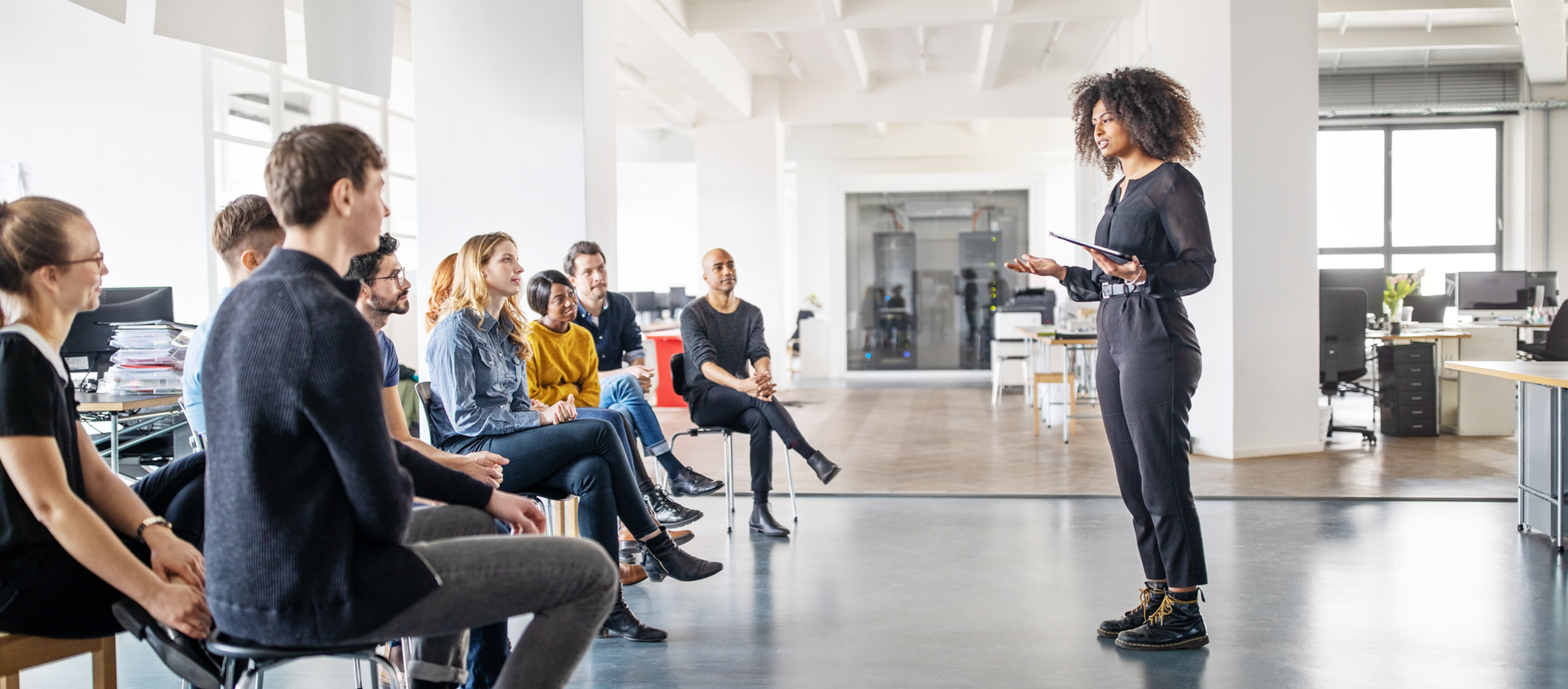 Woman in black speaks to a seated group in a bright open office during a meeting or workshop.