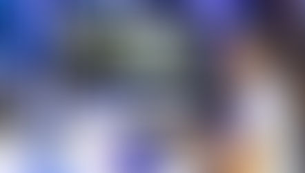 Blurry image of person using a laptop; background shows a wooden setting. Another person sits nearby. Muted colors create a mellow mood.