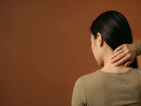Woman seen from behind rubbing her neck against a plain brown background, suggesting tension or pain.