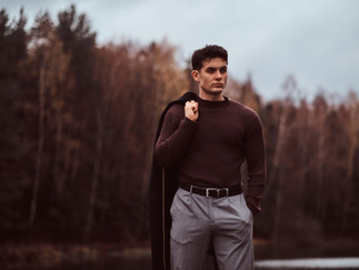 Man wearing long sleeved top and grey trousers while carrying jacket over shoulder for a portrait photo at a forest in autumn