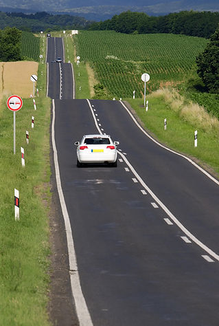 White saloon car driving up a long, winding, undulating road surrounded by fields and crops