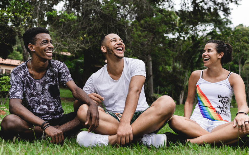 Three young people sitting on grass outdoors laughing together, casually dressed with trees in the background