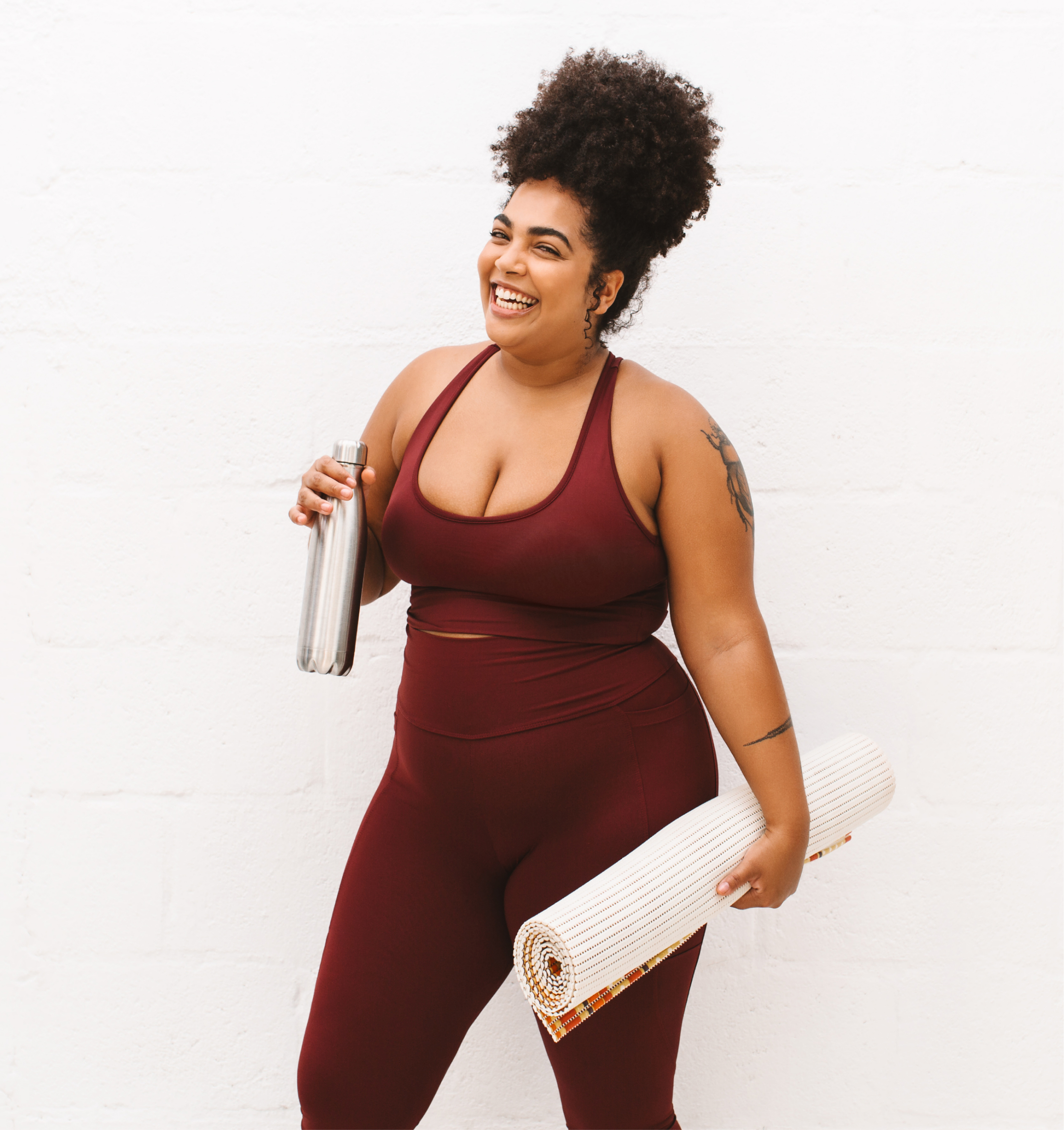 Woman in burgundy activewear holding a yoga mat and water bottle, smiling brightly against a white background.