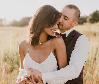 Two people hugging in a field of grains. The female is wearing a white sundress, the male is wearing a white dress and vest