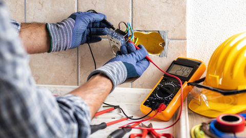 Électricien en gants raccorde une prise murale encastrée, avec multimètre orange affichant 249 et casque jaune à côté.