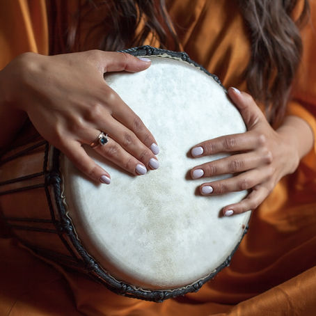 A woman wearing orange clothing, sitting cross-legged, playing a djembe drum on her lap.