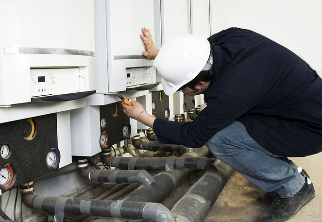 Plumbing engineer in a dark jacket and jeans inspecting a large industrial boiler system