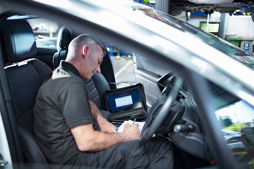 Mechanic sitting in the driver seat writing notes with a diagnostic tablet nearby