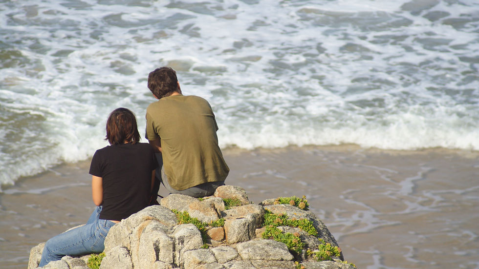 Two people sit on rocky cliffs, facing the ocean waves. One wears a black shirt, the other green. The scene is calm and contemplative.