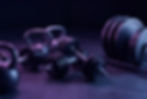 Kettlebells, dumbbells, and weight plates rest on the floor under purple lighting, creating a moody gym atmosphere.