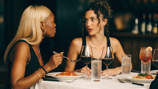 Two women in a dim restaurant talk over pasta, wine and cocktails at a white-clothed table.