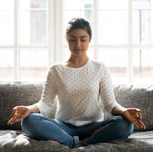 Woman sitting cross-legged on a gray sofa, practicing meditation to build somatic awareness in a calm, sunlit living space.