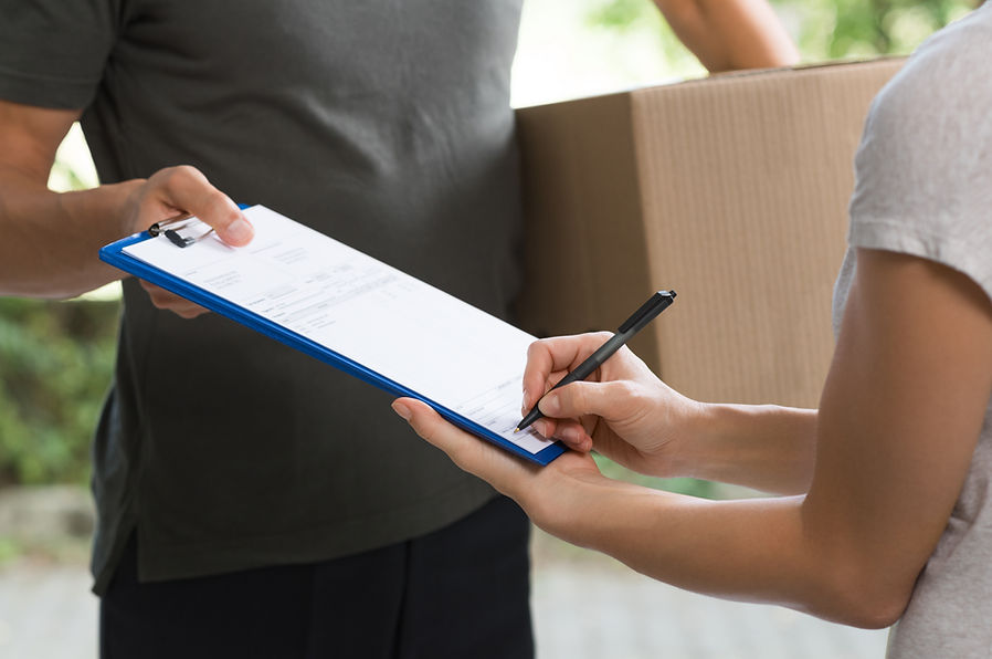 A person signing a delivery receipt while holding a clipboard, with another individual holding a cardboard box