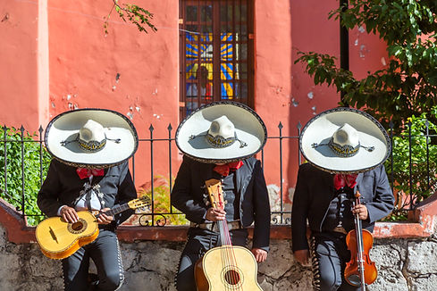 Mariachi musicians in traditional black suits and wide-brimmed sombreros standing against a red stucco wall.